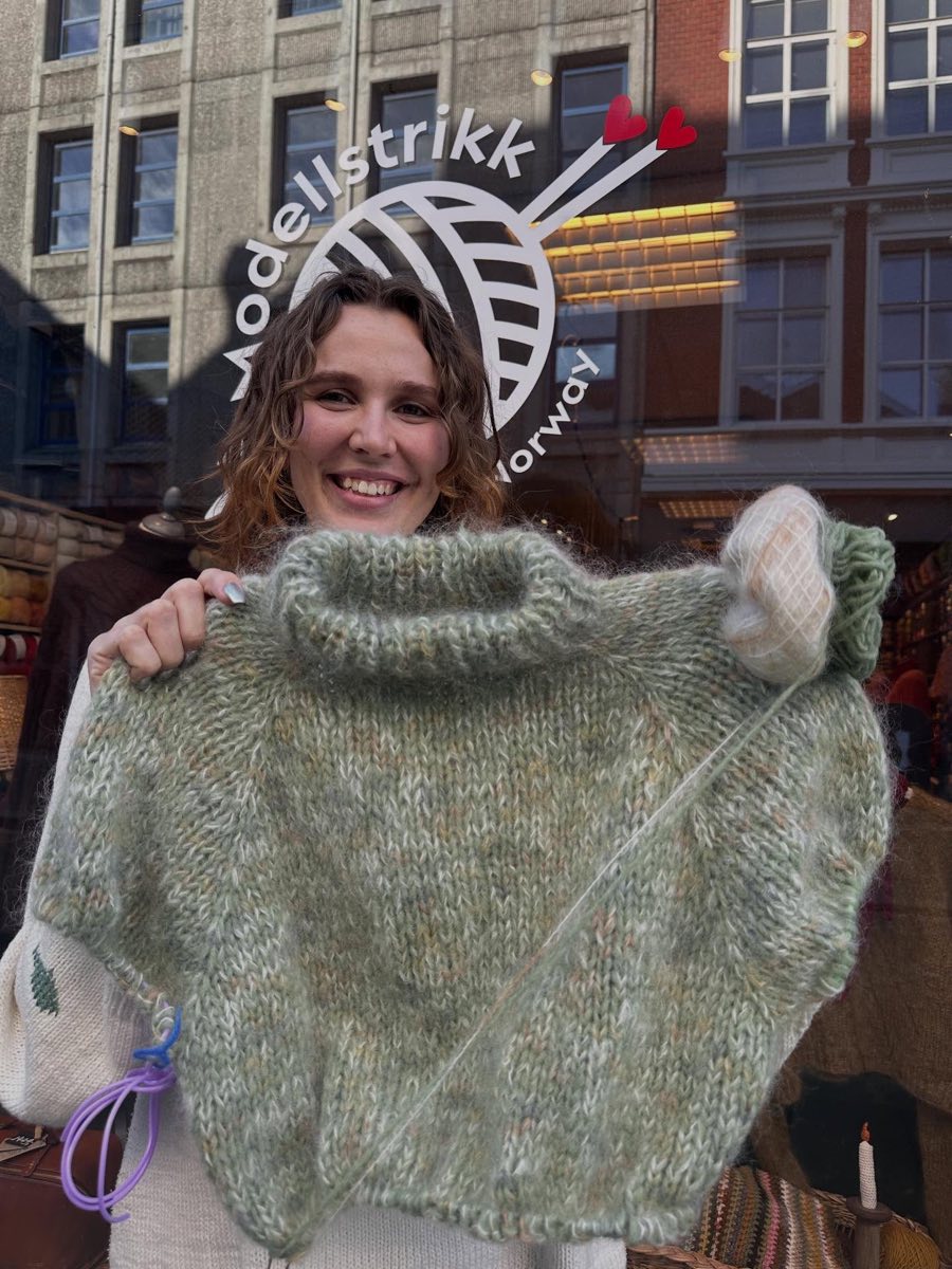 Charlotte holder opp en strikket genser foran butikkvinduet med Modellstrikk-logoen